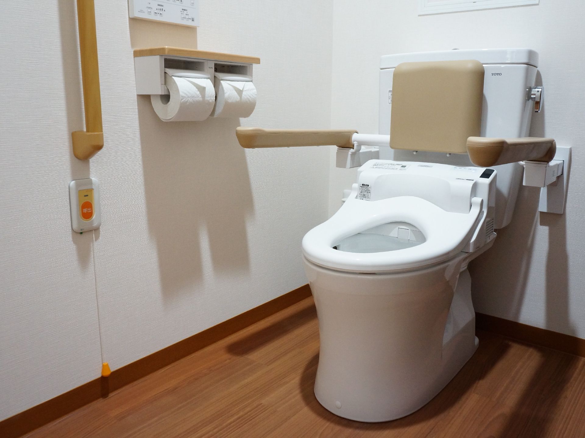 壁に手すりとナースコールが付いている。便器の両脇にも可動式の手すりが設置されているほか、背当ても付いている。