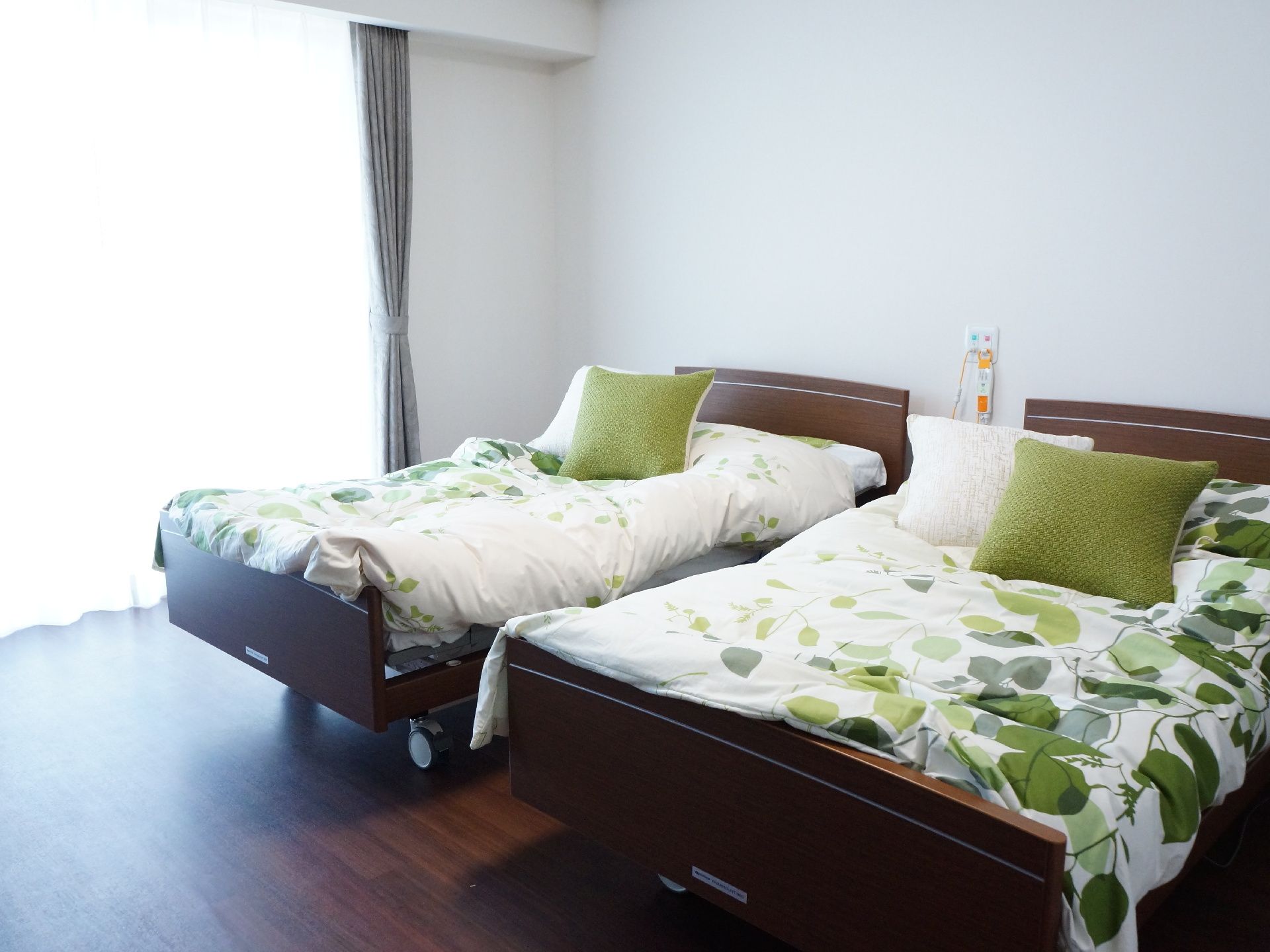 大きな掃き出し窓のある明るい室内に、介護ベッドが二台並んで置かれている。部屋は白壁にダークブラウンのフローリングである。