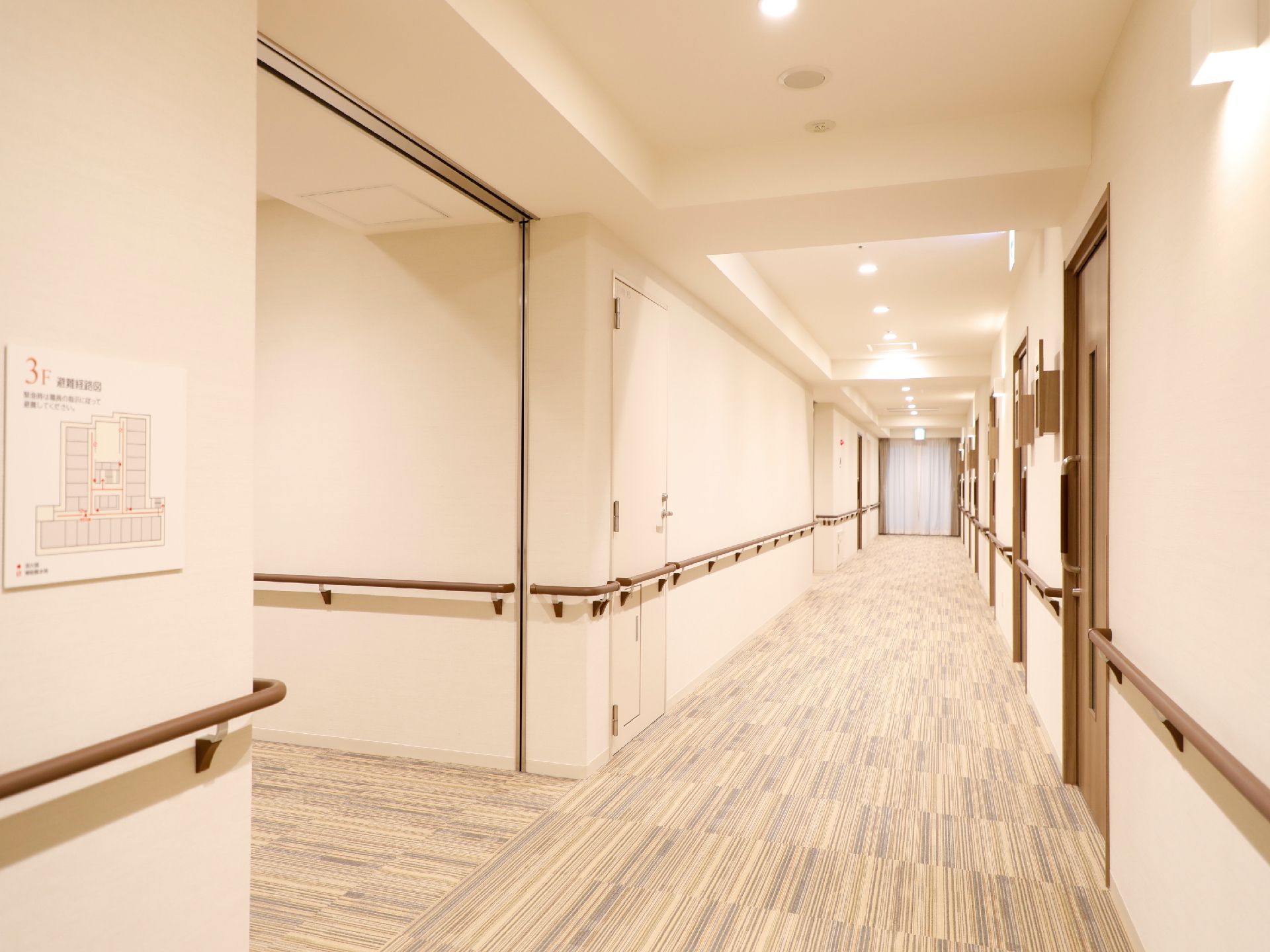 左手前の壁に、施設3階の案内図が掲示されている。廊下の壁は白色で、ダークブラウンの手すりが設置されている。