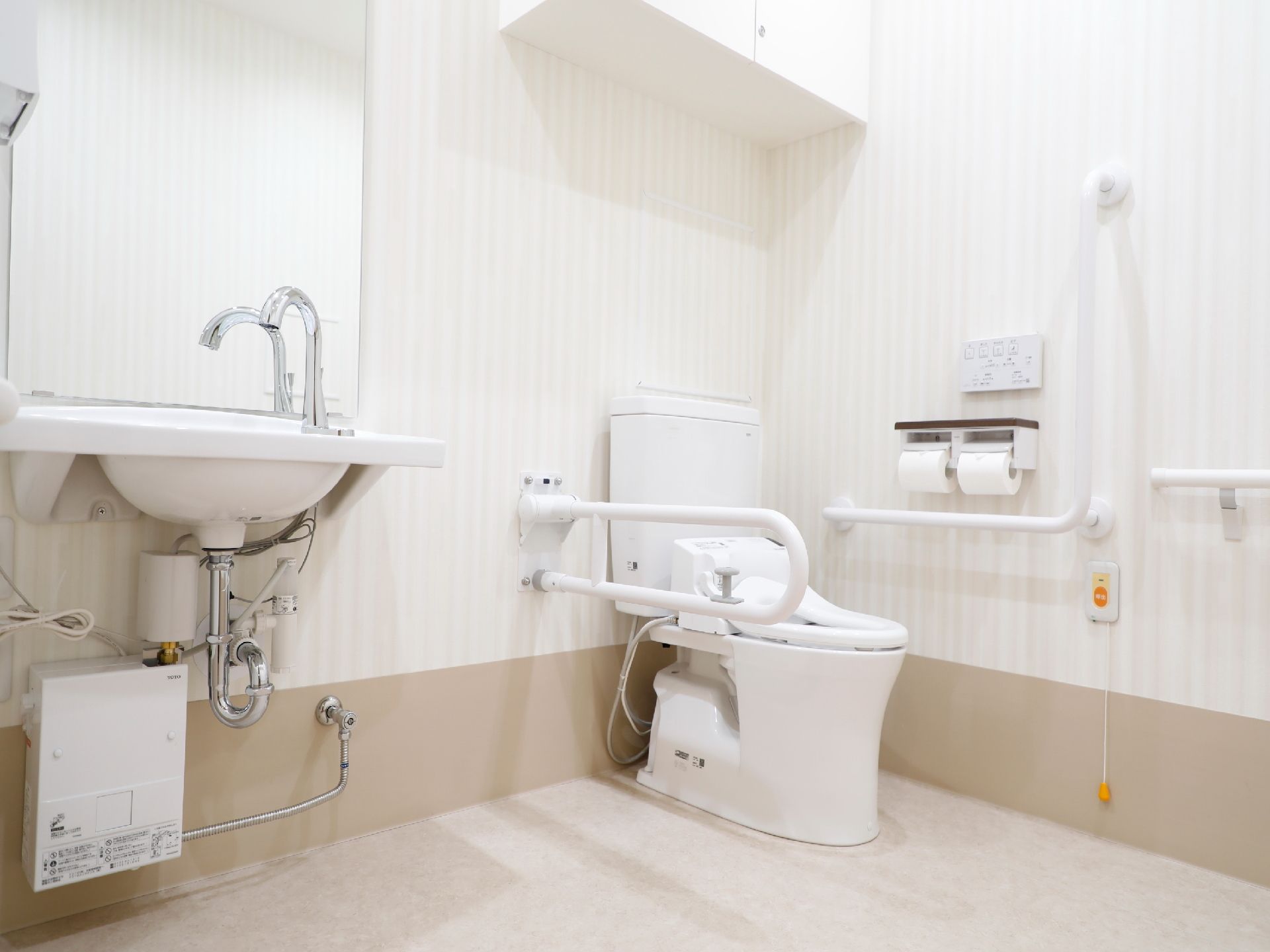 車椅子仕様のトイレである。可動式やL字型の手すり、呼び出しボタンが壁に取り付けられている。洗面台も設置されている。