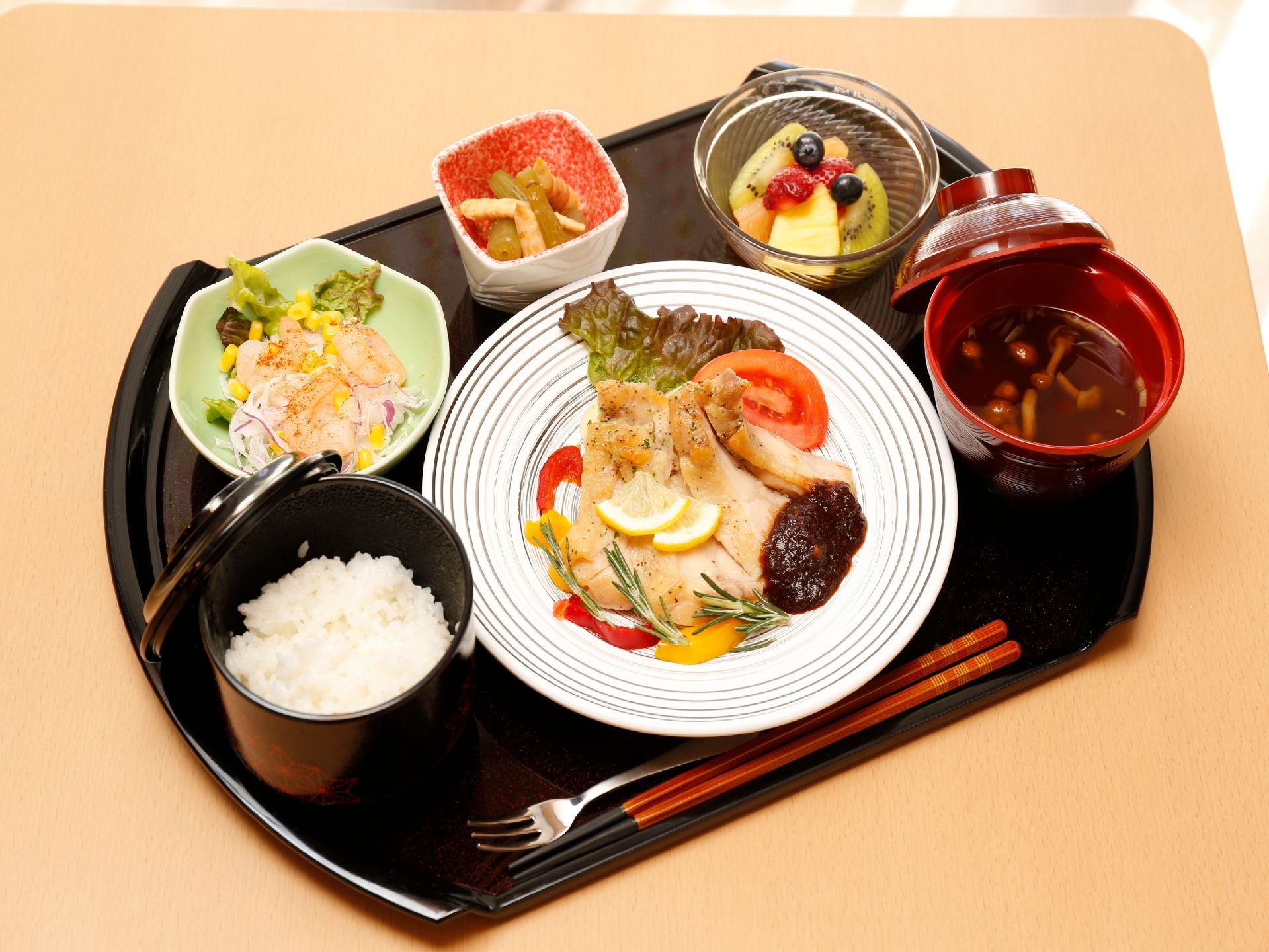 施設で出される食事の例である。メインの鶏肉料理とサラダ、小鉢、ご飯、みそ汁、デザートの果物など品数豊富だ。