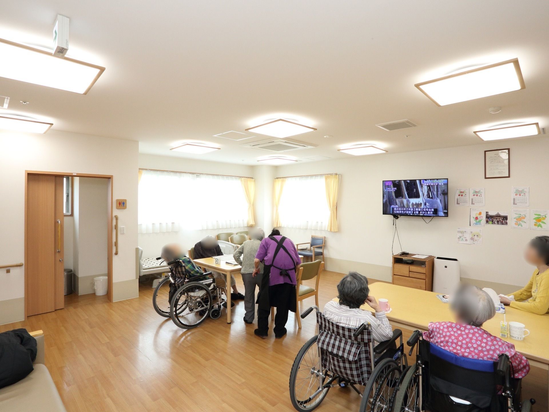 テーブルと椅子が並べられ、入居者様が座っている。壁にはテレビが掛けられており、奥に共用トイレが設置されている。