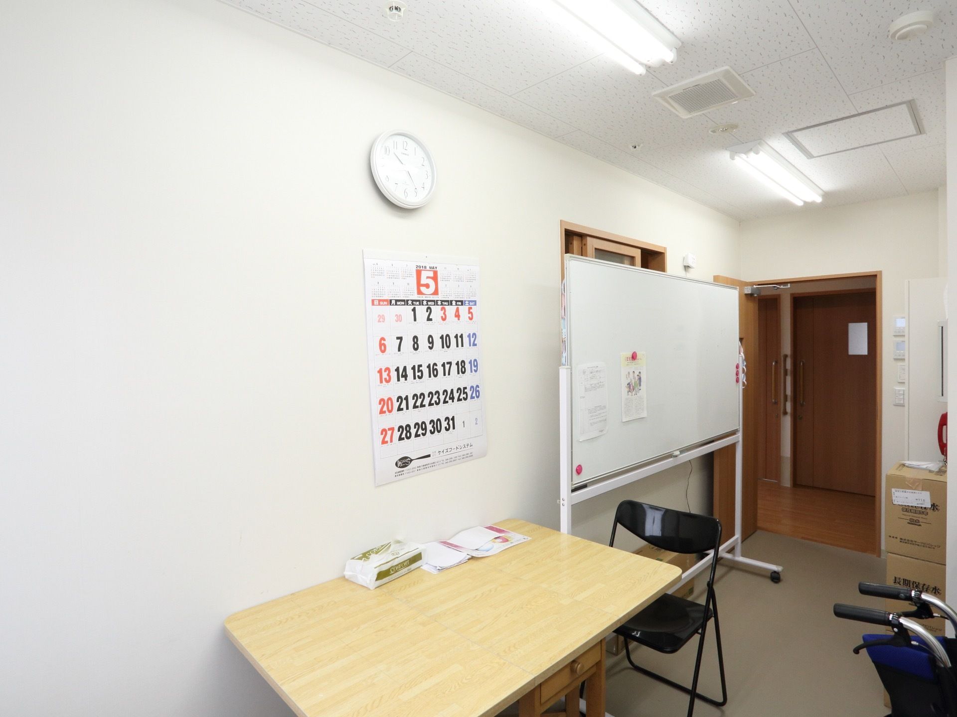 テーブルと椅子が壁に向かって設置されており、入り口付近にホワイトボードが置かれている。壁にはカレンダーと白い時計が掛けられている。