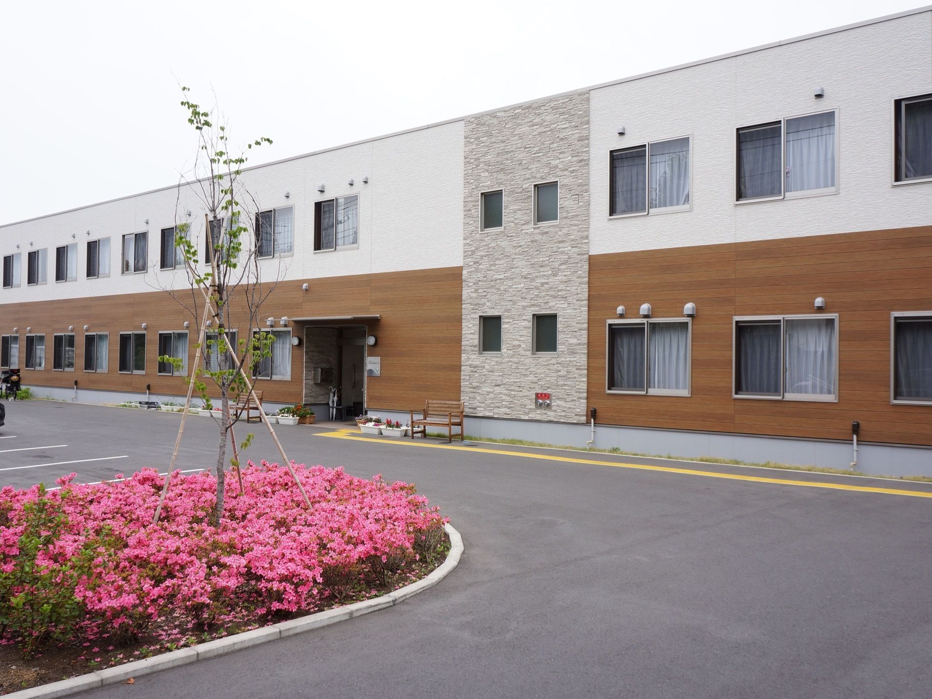 ２階建てで、フロアごとに外壁が塗り分けられている。施設前の樹木の植え込みはピンクの花に包まれている。