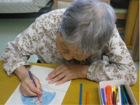 色鉛筆で白い紙にうちわの絵を描いている、白髪のおばあさん