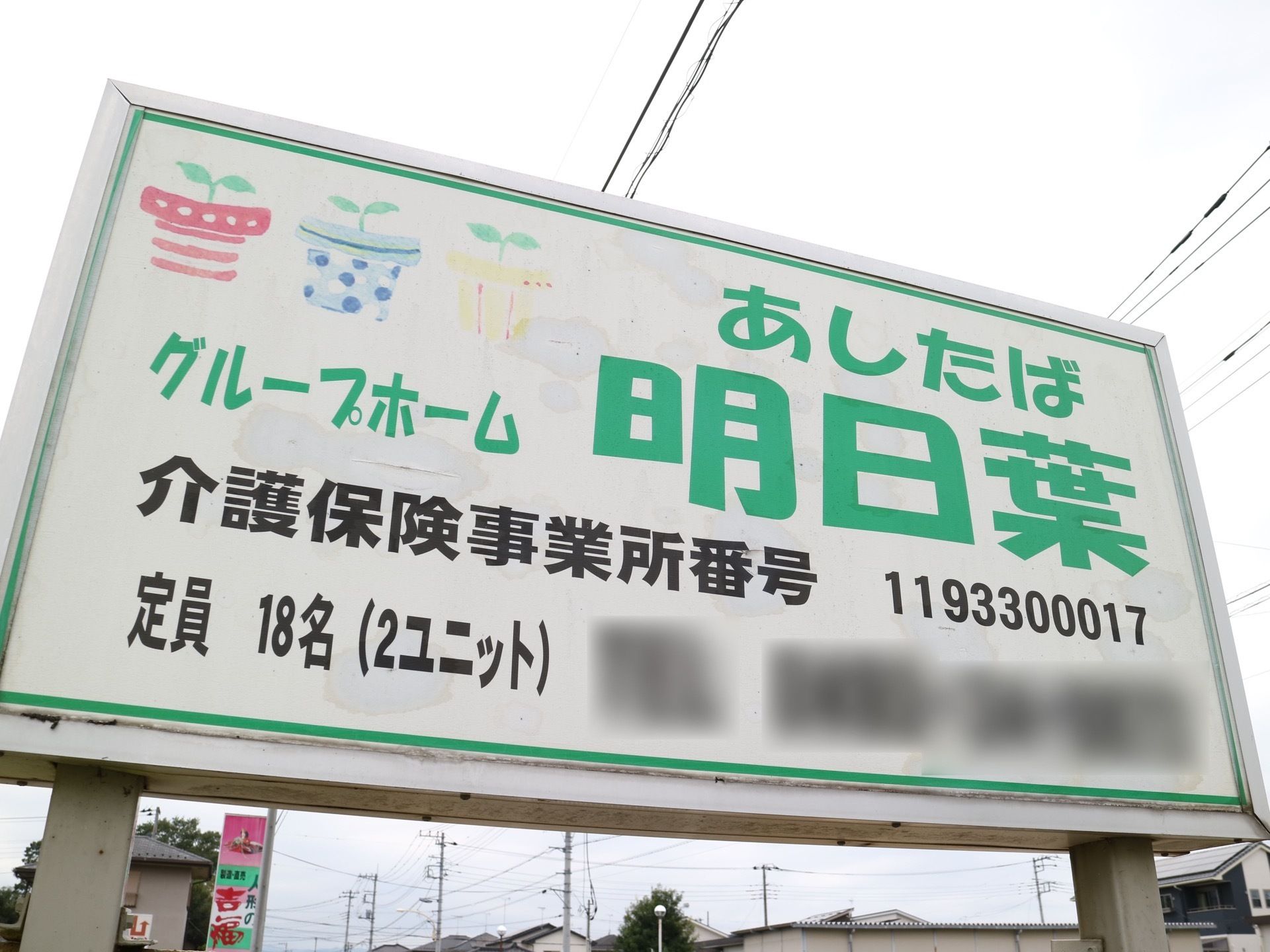 支柱の上の高い位置に横長の看板が取り付けられている。施設名称の漢字と読み仮名が大きな緑色の文字で表示されている。