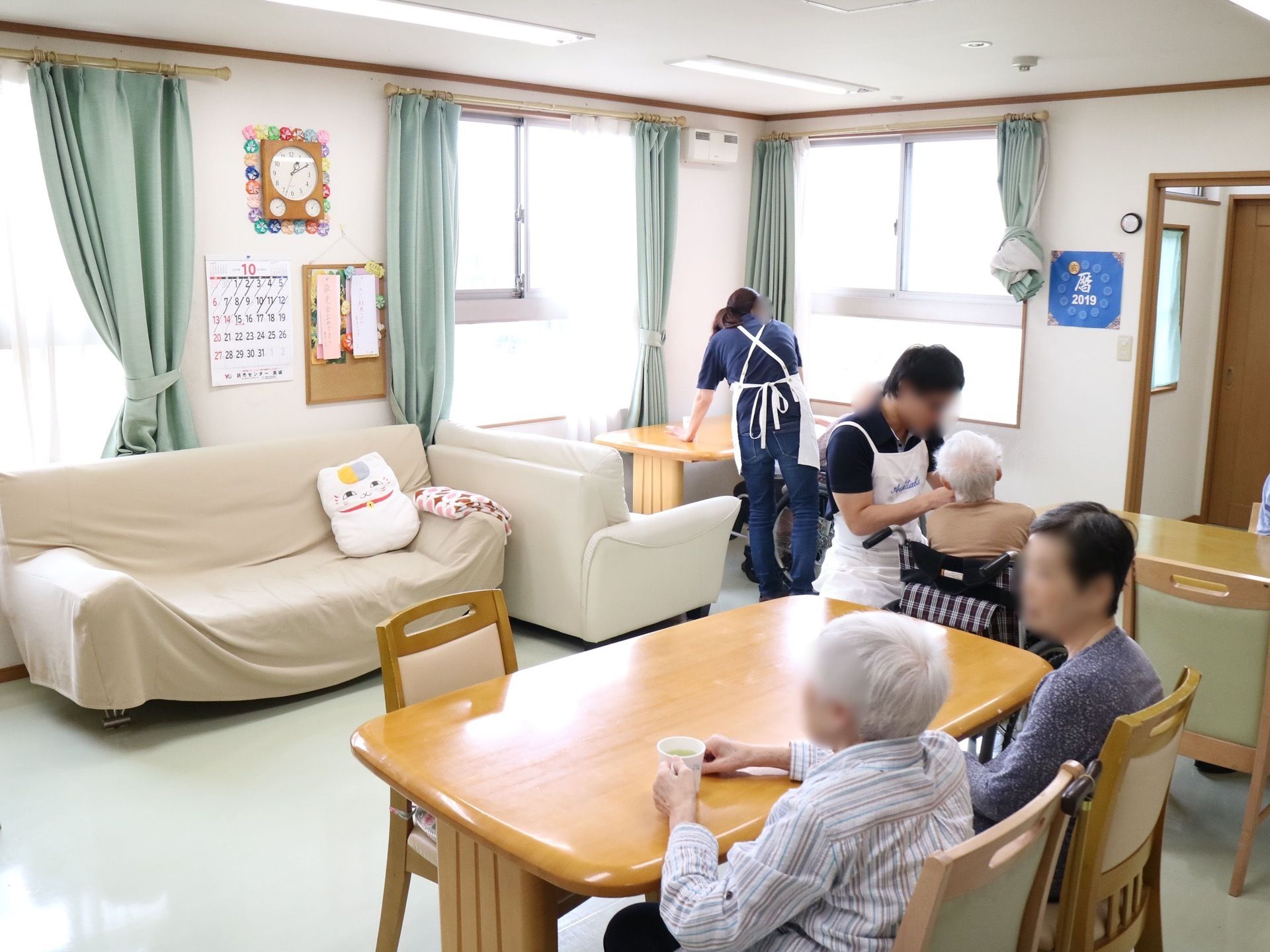 テーブルや椅子、ソファが置かれた共有スペースがある。車椅子をご利用の入居者様の近くに、エプロンをしたスタッフが立っている。