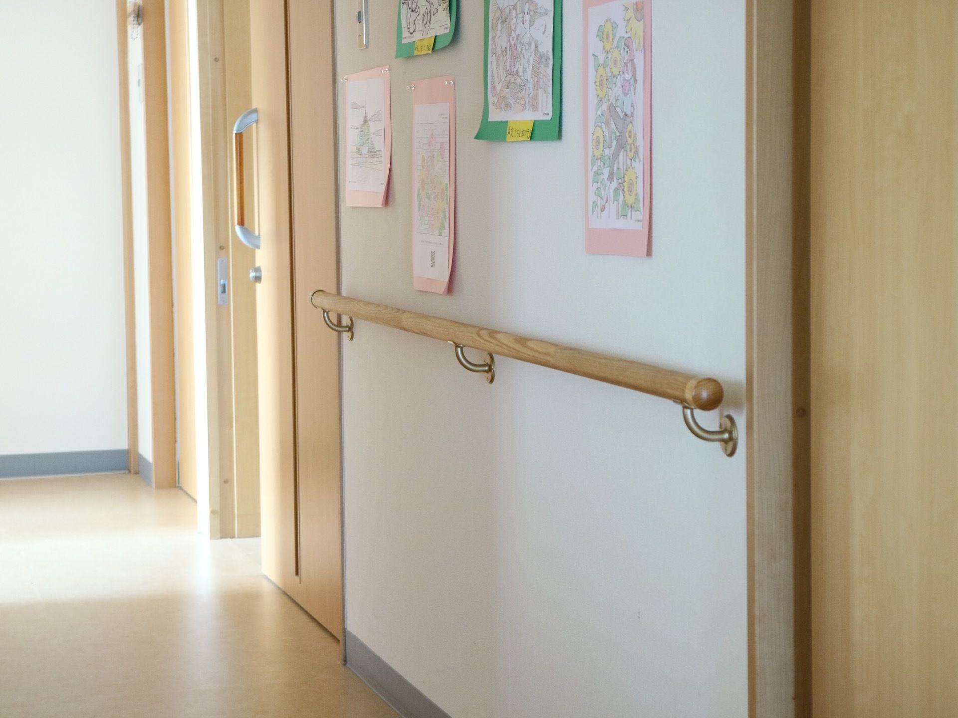 入居者様の絵が飾られた廊下がある。壁には木目調の手すりがある。居室やトイレの入口も木目調の扉が付いている。