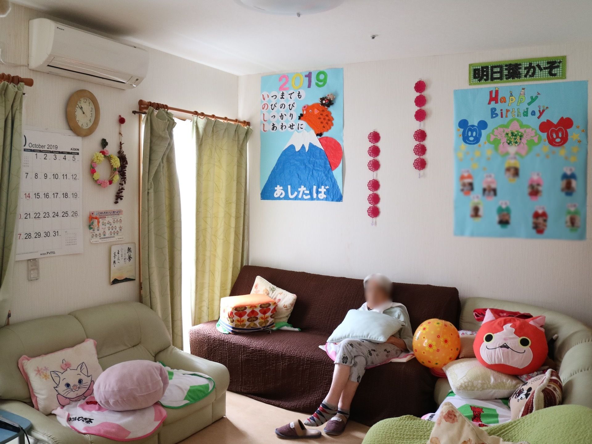 ソファがたくさん置かれたスペースがある。掃き出し窓があり、壁にはエアコンがある。入居者様が１人、腰を掛けて寛いでいる。