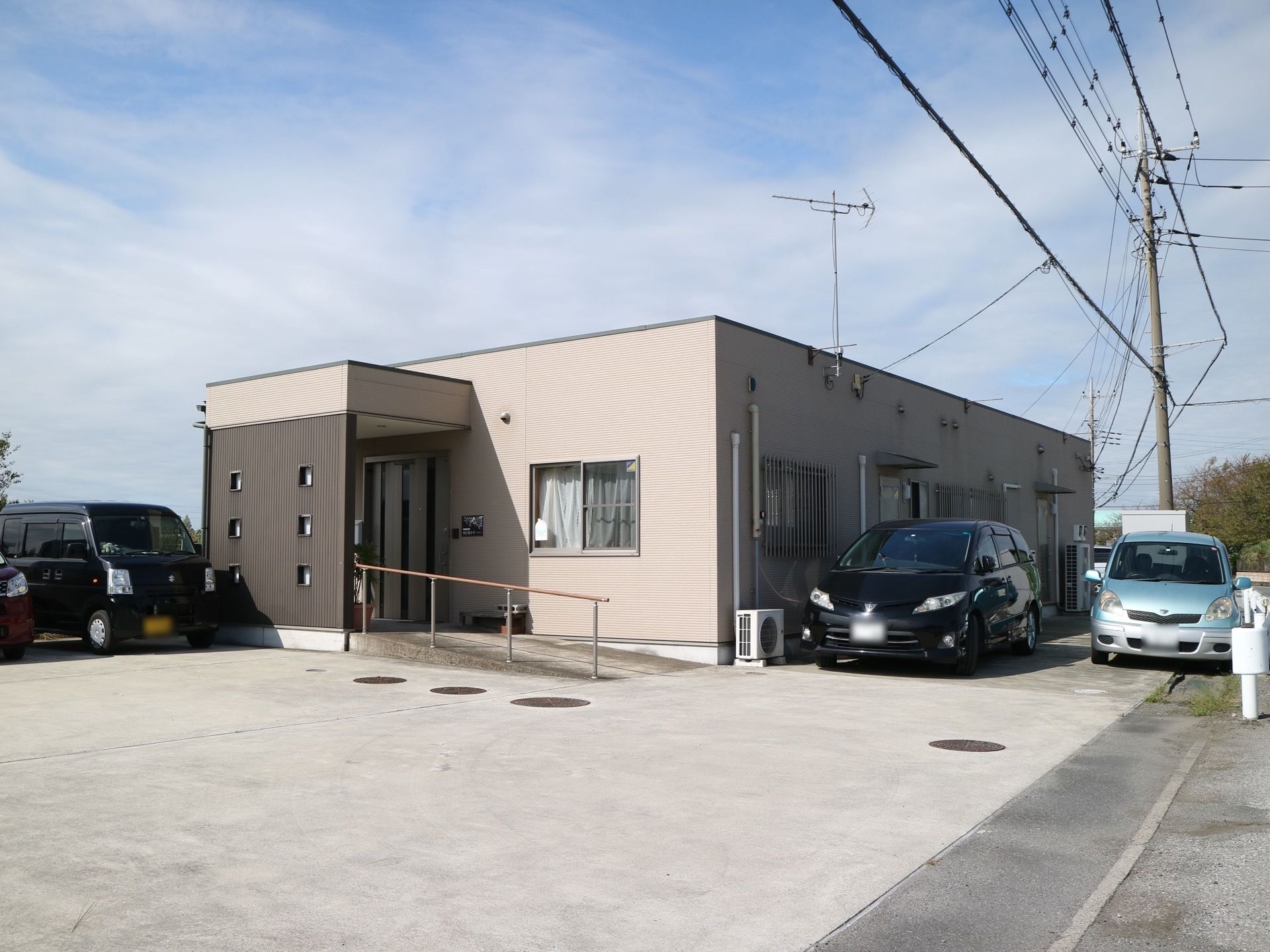落ち着いたグレー調の施設外観である。平屋建てになっており、駐車スペースには車が何台か停められている。