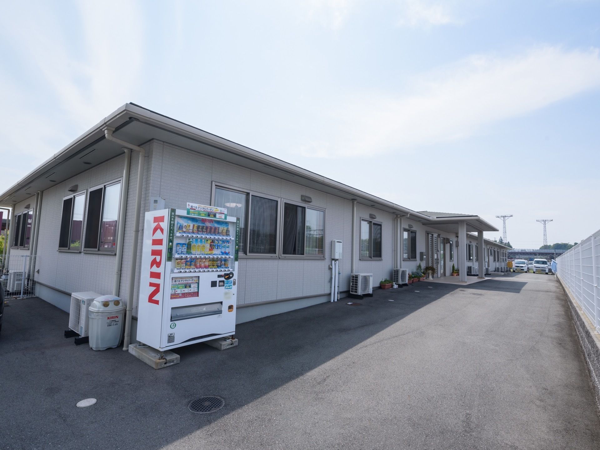 施設の建物は淡いグレーを基調とした平屋である。建物に沿ってエアコンの室外機や自動販売機が設置されている。