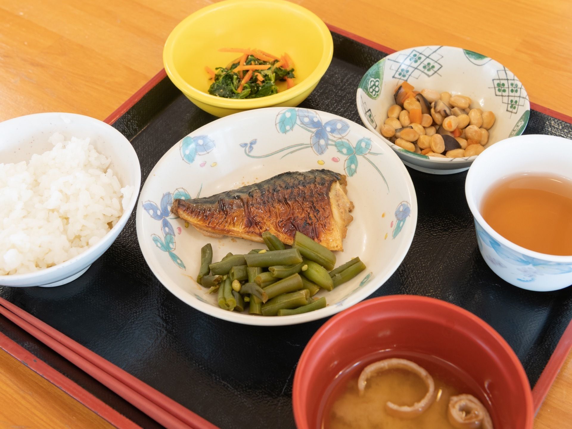 縁が赤色に彩られた、お盆に料理が置かれている。今日の料理は魚がメイン。白ご飯にお味噌汁、小鉢もある。
