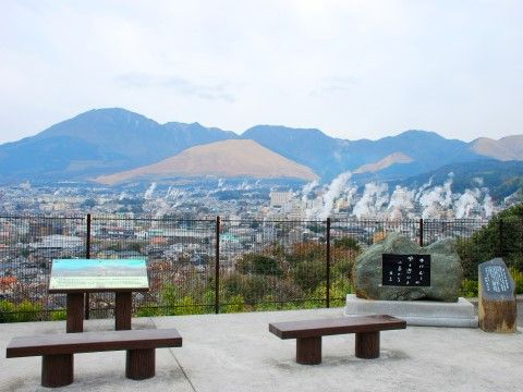 展望台のような場所にベンチが2つあり、石碑などが設置されている。遠くに山が見え、その手前のあちこちに湯けむりが立ち上っている。