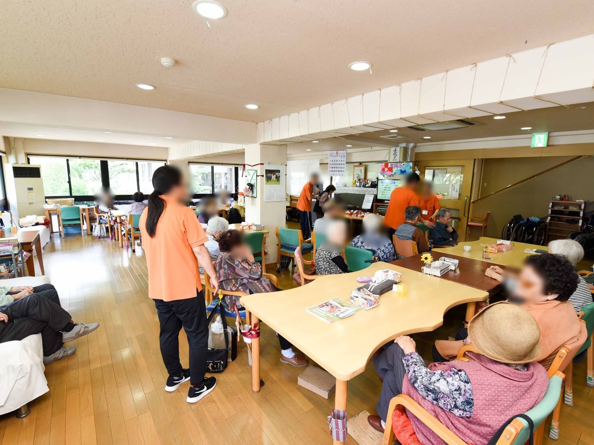 テーブルと椅子が設置され、たくさんの入居者様が座っている。オレンジのポロシャツを着たスタッフが、入居者様に寄り添いサポートしている。