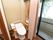 窓の近くにトイレが設置されている。便座には木目調の手すりが付いており、床はフローリングになっている。