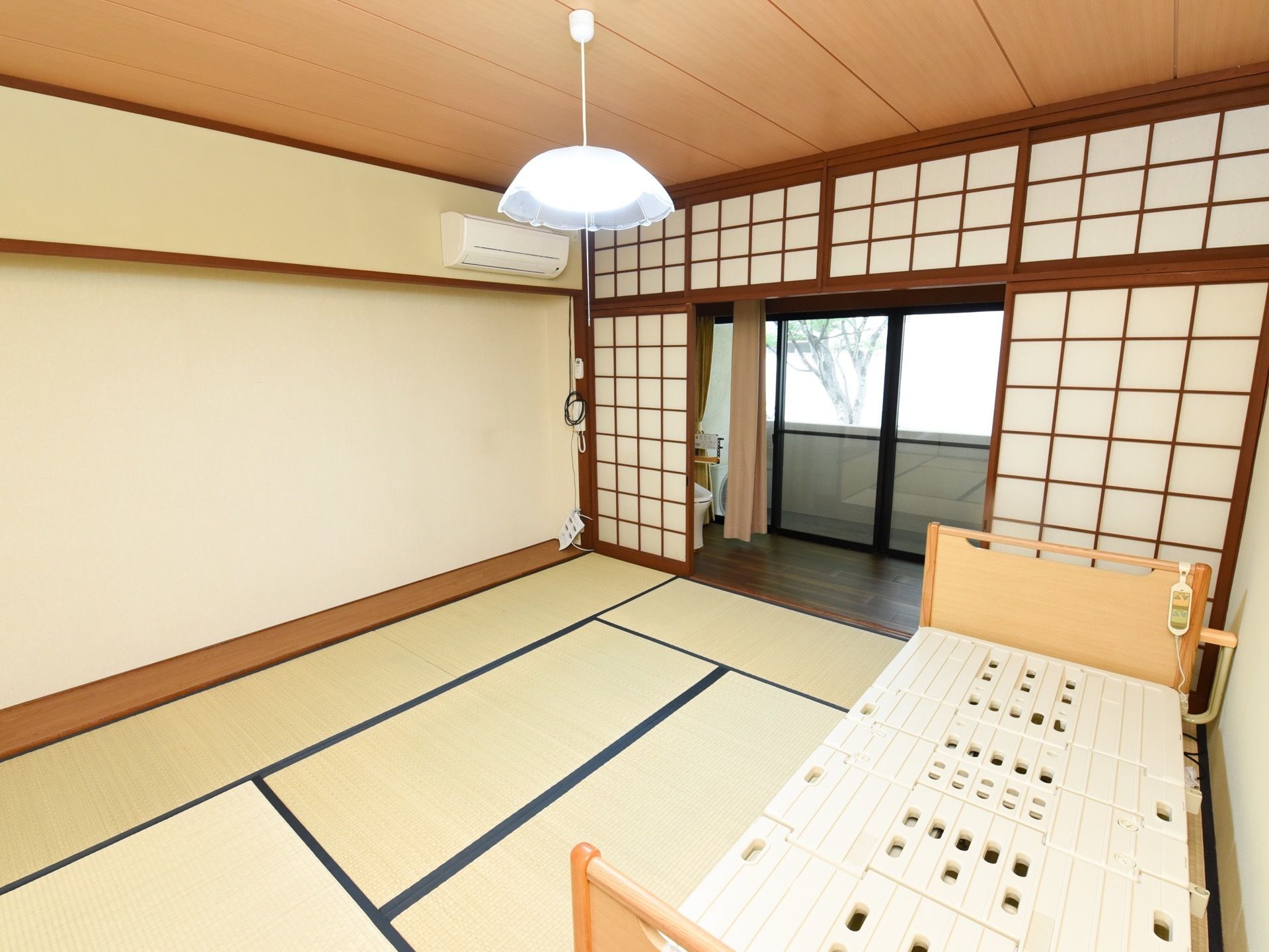 畳の部屋に電動ベッドが置かれている。障子の向こうにはトイレが設置されており、カーテンが付けられている。