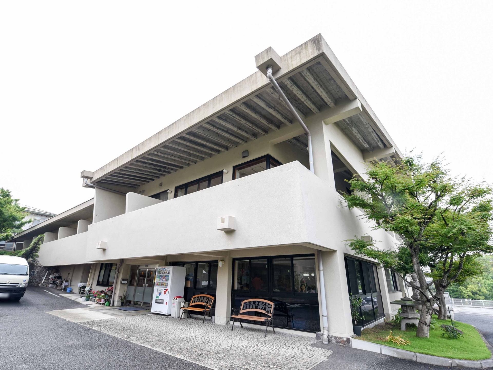 白い外壁の2階建てになっている。1階部分には大きな窓があり、その前にはベンチや自動販売機が設置されている。