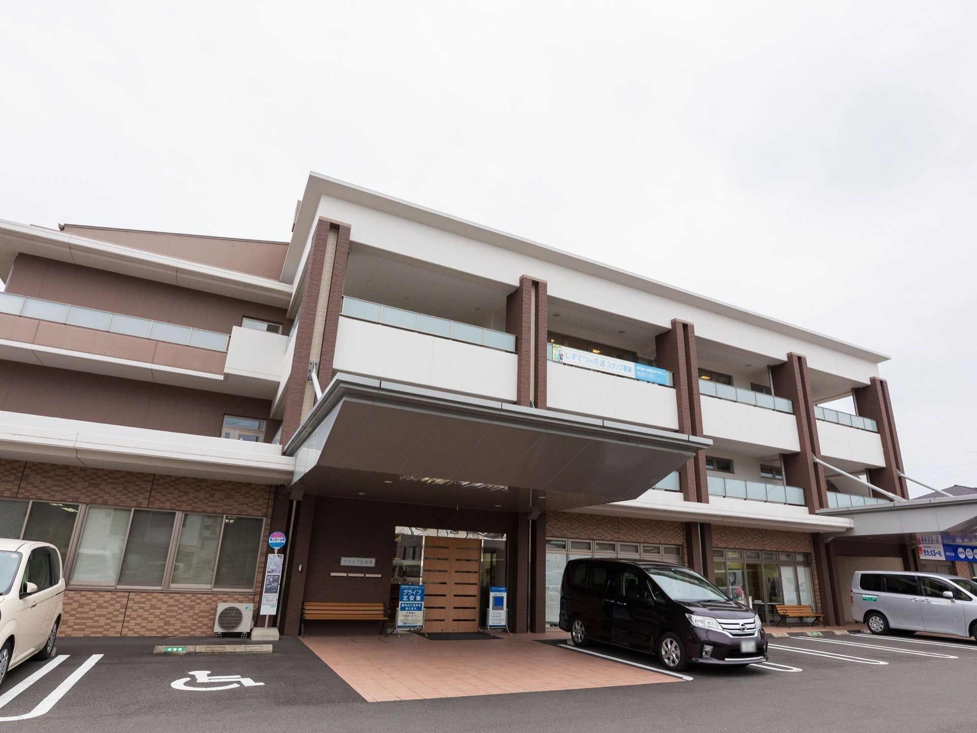 施設の建物は３階建てで、白色と茶色を基調としている。１階の自動ドアの前には大きな屋根があり、隣には身障者用の駐車スペースが設けられている。