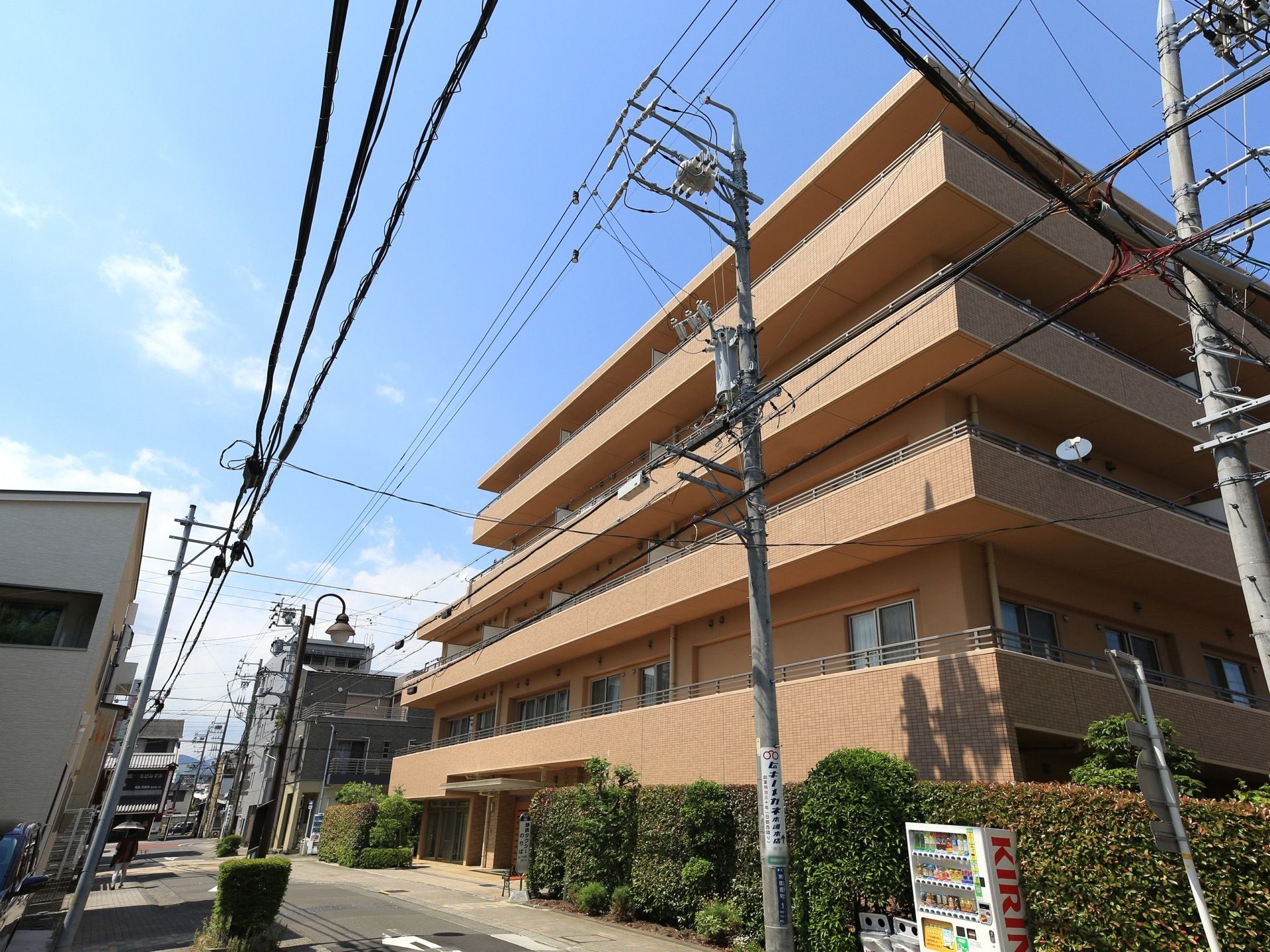 茶色い外壁の５階建ての建物で、建物の周囲には植物が植えられている。手前に白い自動販売機が設置されている。