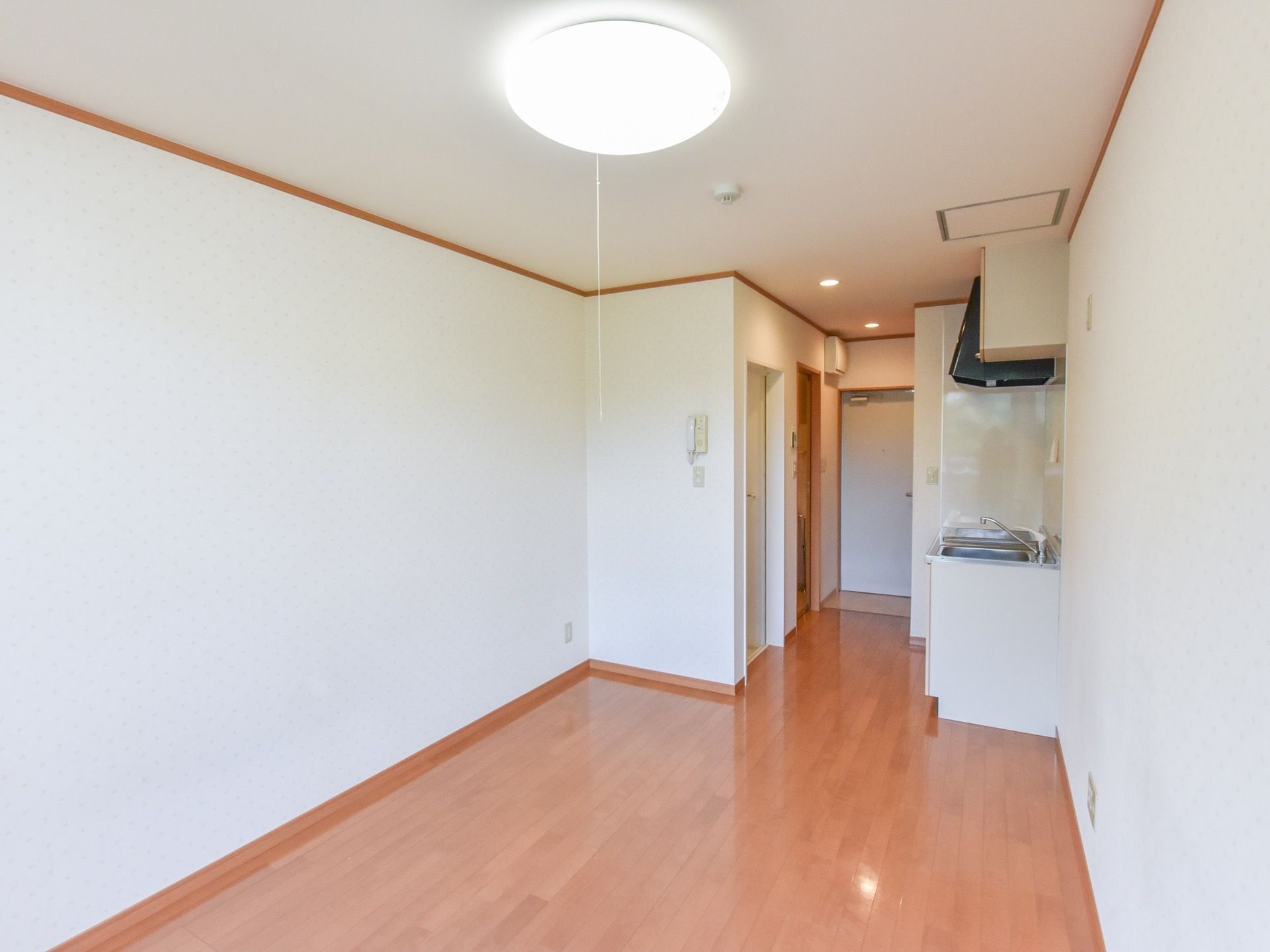 白い壁にフローリングの、ワンルームタイプの部屋。入口を入って左側にミニキッチンが備え付けられている。