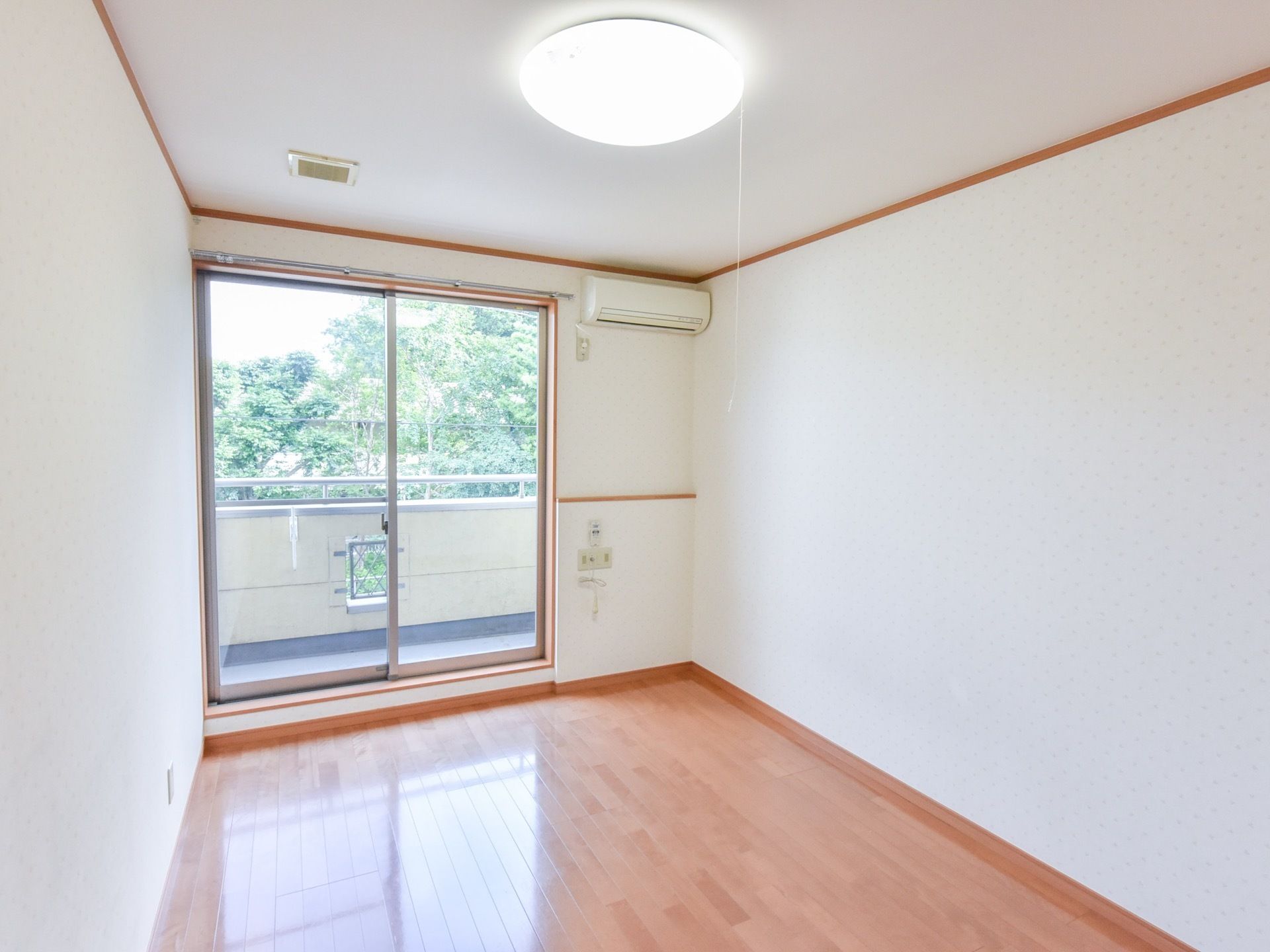 ベランダへ出て外の空気が楽しめる明るい居室。エアコンがついている。窓の外には、緑の生い茂った木々が見える。