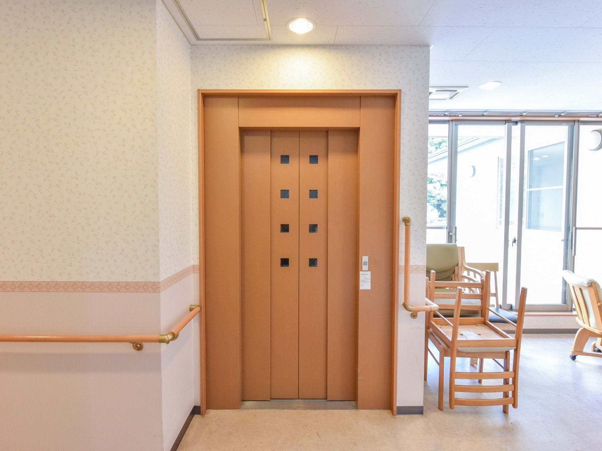 エレベーターが設置されている。木目調のあたたかい雰囲気の扉である。周りの壁には補助用の手すりがついている。