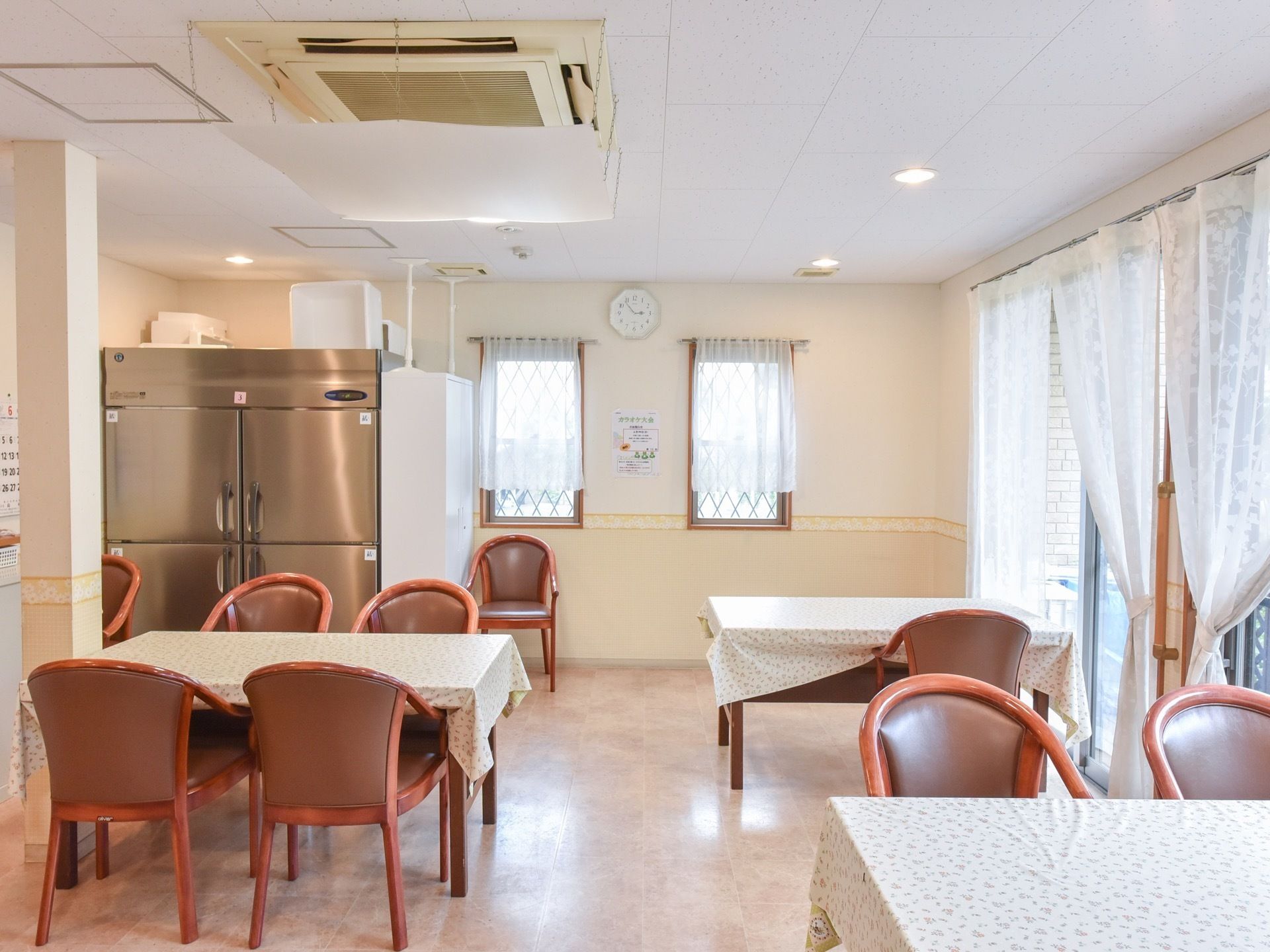 食堂の奥には、業務用の大きな冷蔵庫や棚が設置されている。手前には食事用のテーブルが並べられている。テーブル同士の間隔は広く取られている。