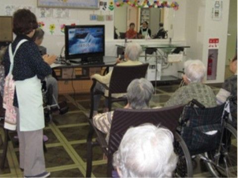 椅子に座ってテレビを見ている数名の高齢者とエプロンをつけている女性