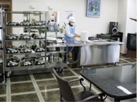 ステンレス製の調理台で、白い帽子とマスクを身に付けた水色のエプロン姿の人物が作業をしている。壁に絵画が飾られている。