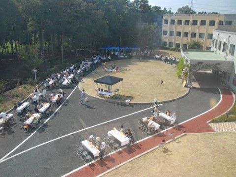 建物の前のロータリーにたくさんのテーブルと椅子が並べられ、テーブルの周りに多くの人が座ったり、立ったりしている。
