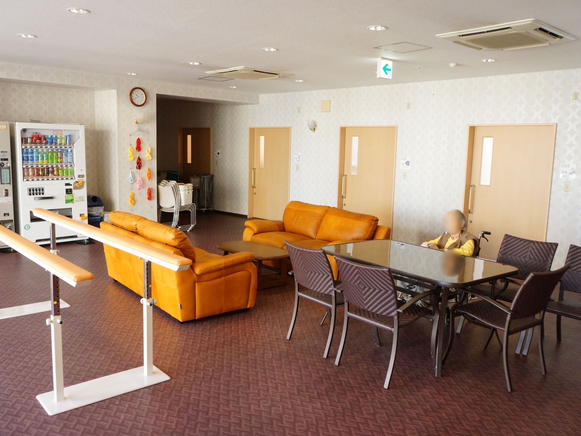 広々とした休憩スペースには、平行棒やソファーが置いてある。左端には自販機があり、その横の壁には時計が掛けてある。