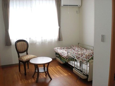 電動リクライニング式ベッドを設置している。窓からは日差しが差し込み明るい室内。窓際には椅子とテーブルが置いてある。