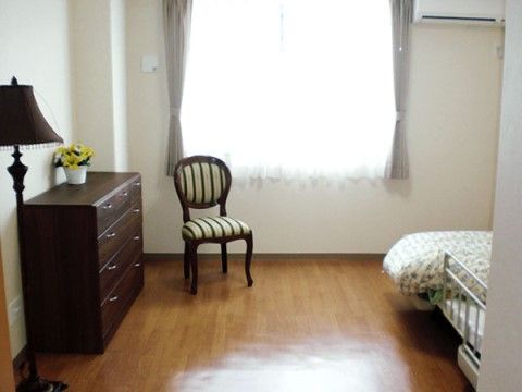 壁際に４段のチェストがあり、その上には黄色い花が飾られている。近くにはスタンドライトと椅子が１脚置かれている。