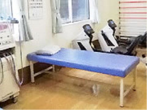 白い壁にフローリングの床の室内。リハビリに使用するブルーの簡易ベッドが置かれている。ベッドの上には枕がある。