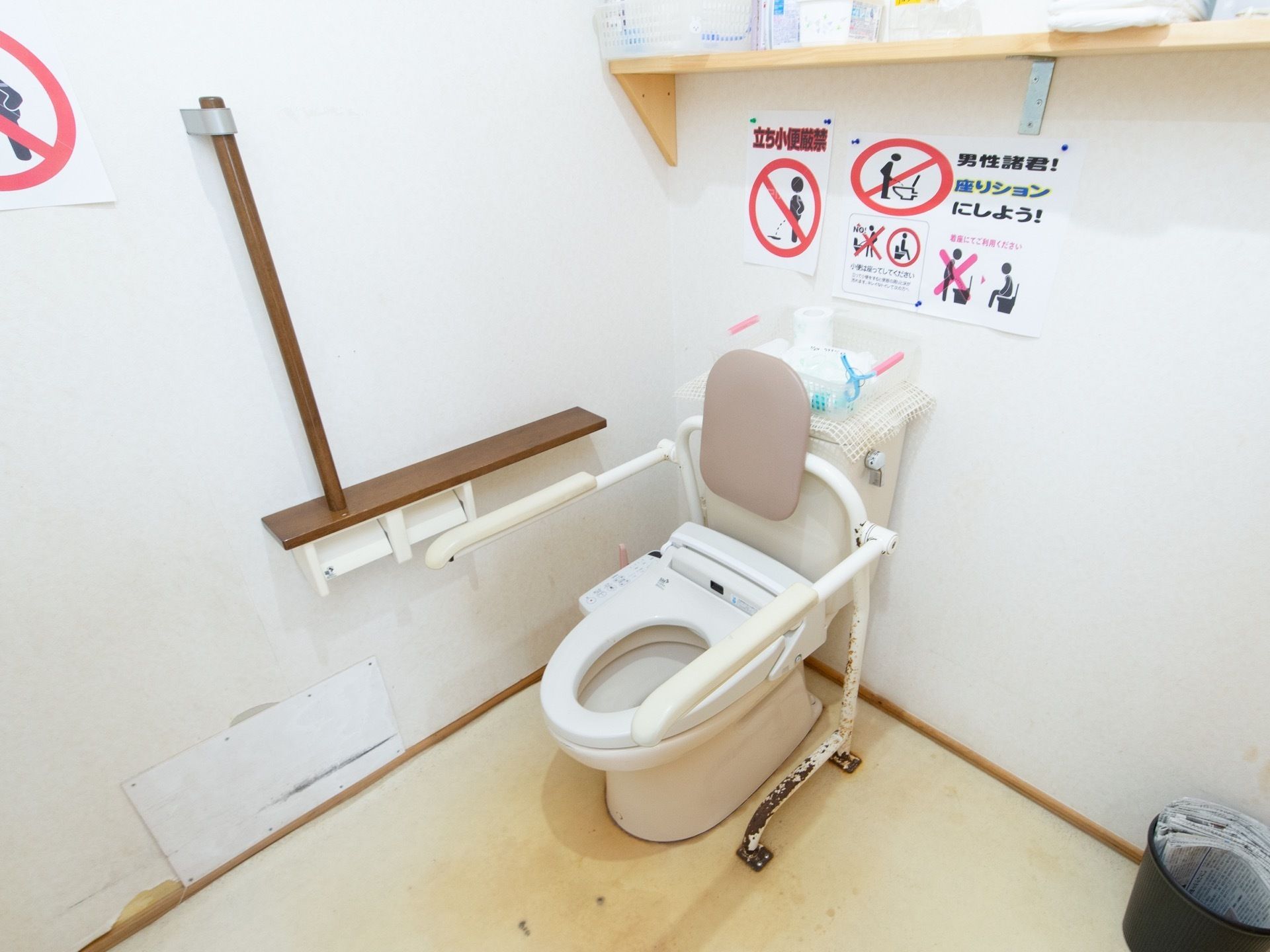 トイレには蓋がなく、クッション性あるひじ掛けと背もたれが付いている。壁には貼り紙がされている。木製の手すりが設置されている。