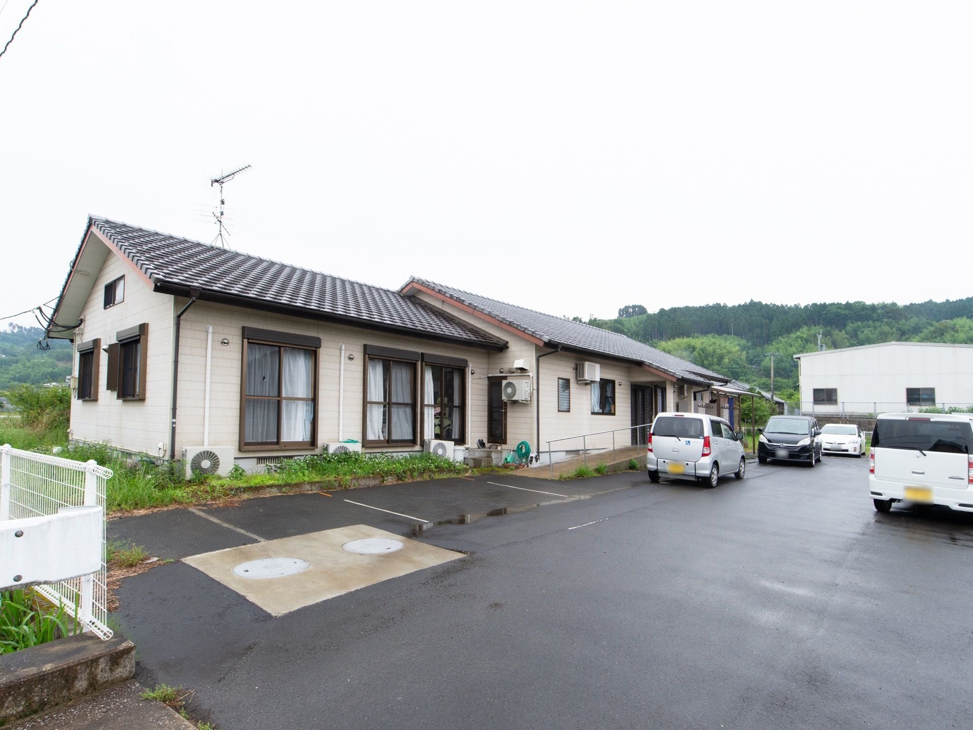 平屋建ての施設があり、周囲は緑に囲まれている。建物のそばには広々とした駐車場があり、車両が数台とめられている。