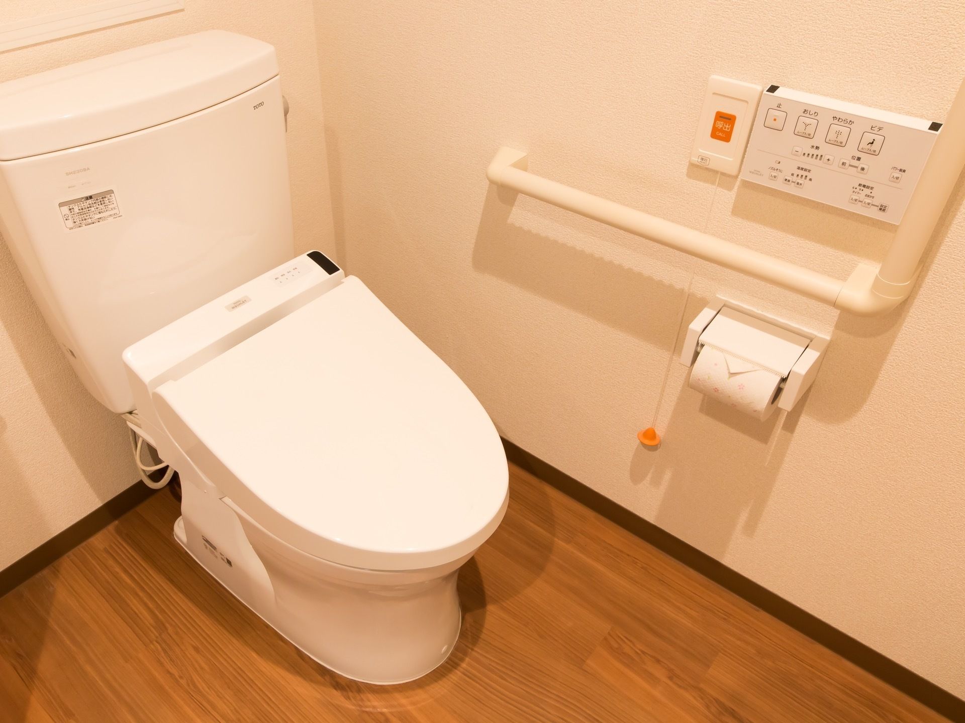 トイレはふたつきの便器である。壁に設置されたＬ字型の手すりの脇に、洗浄暖房機能の操作パネルが取り付けられている。