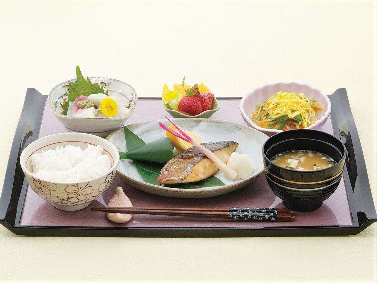黒と紫色のお盆の上に、ご飯、お味噌汁、焼き魚、お刺身、フルーツなどが沢山載っている