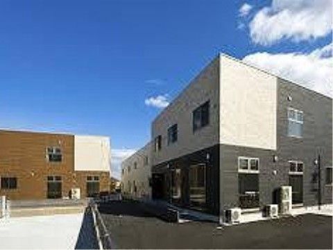 手前には外壁が白とグレーの２階建ての建物があり、その奥には白い外壁の建物の一部が見える。道を挟んで左奥に外壁が白と茶の２階建ての建物がある。