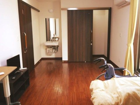 白と木目調を基調とした居室である。壁には鏡付きの洗面台やエアコンが取り付けられている。壁側にはテレビとテーブル、窓側にはベッドが置いてある。
