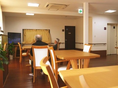 四角いテーブルと背もたれが高い椅子のダイニングセットが複数置いてある共有スペースである。入居者様がテレビを鑑賞している。