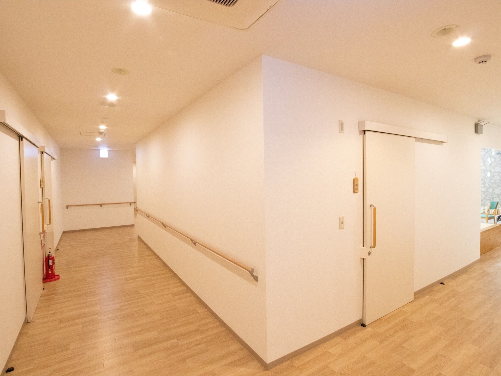 廊下の壁は白で、床に消火器が置かれている。天井に等間隔に照明が埋められている。壁に手すりが設置されている。