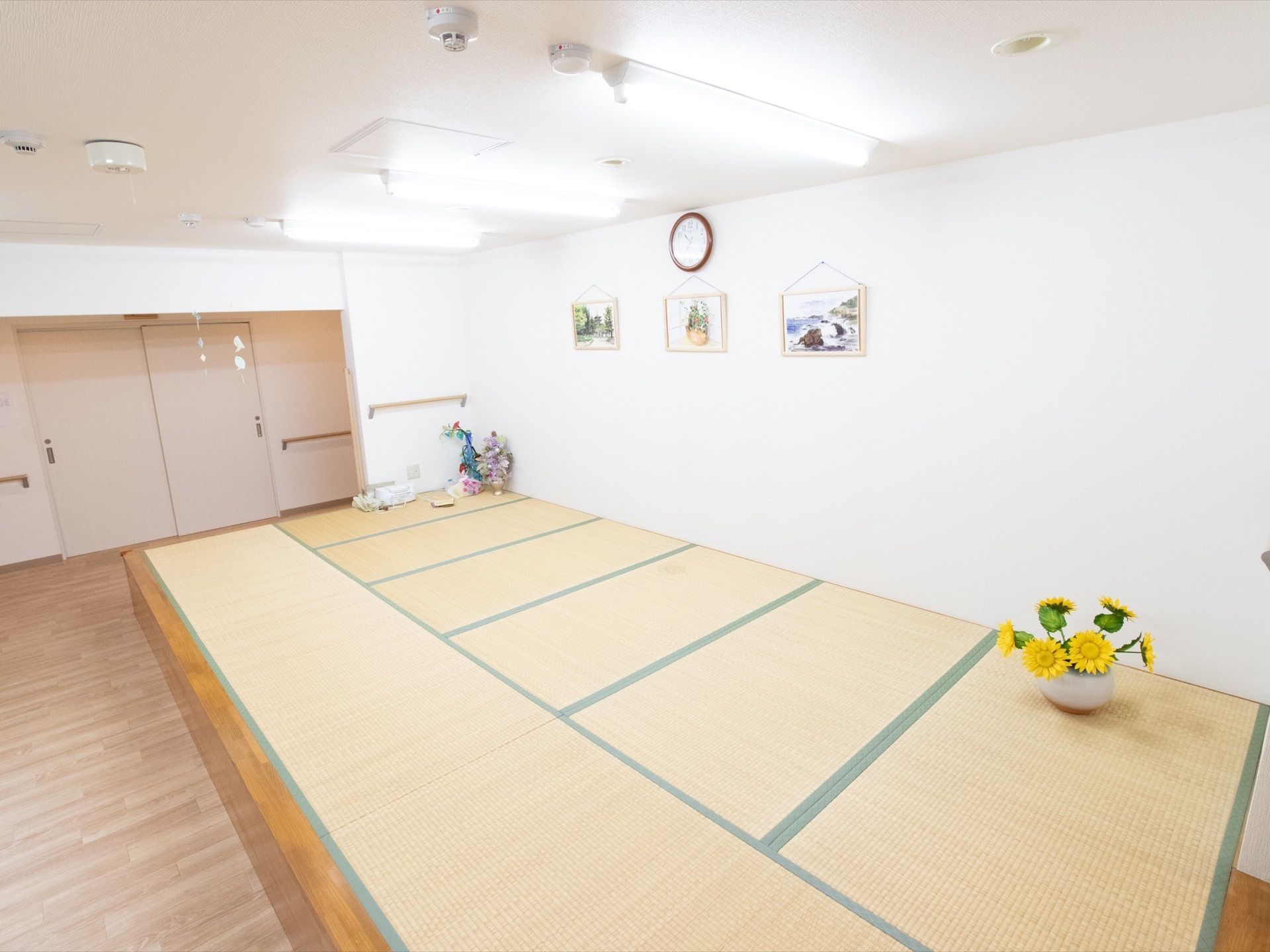 畳の敷かれた休憩スペースがある。壁に絵画が３か所に飾られ、時計が下がっている。床に花瓶にひまわりが生けてある。