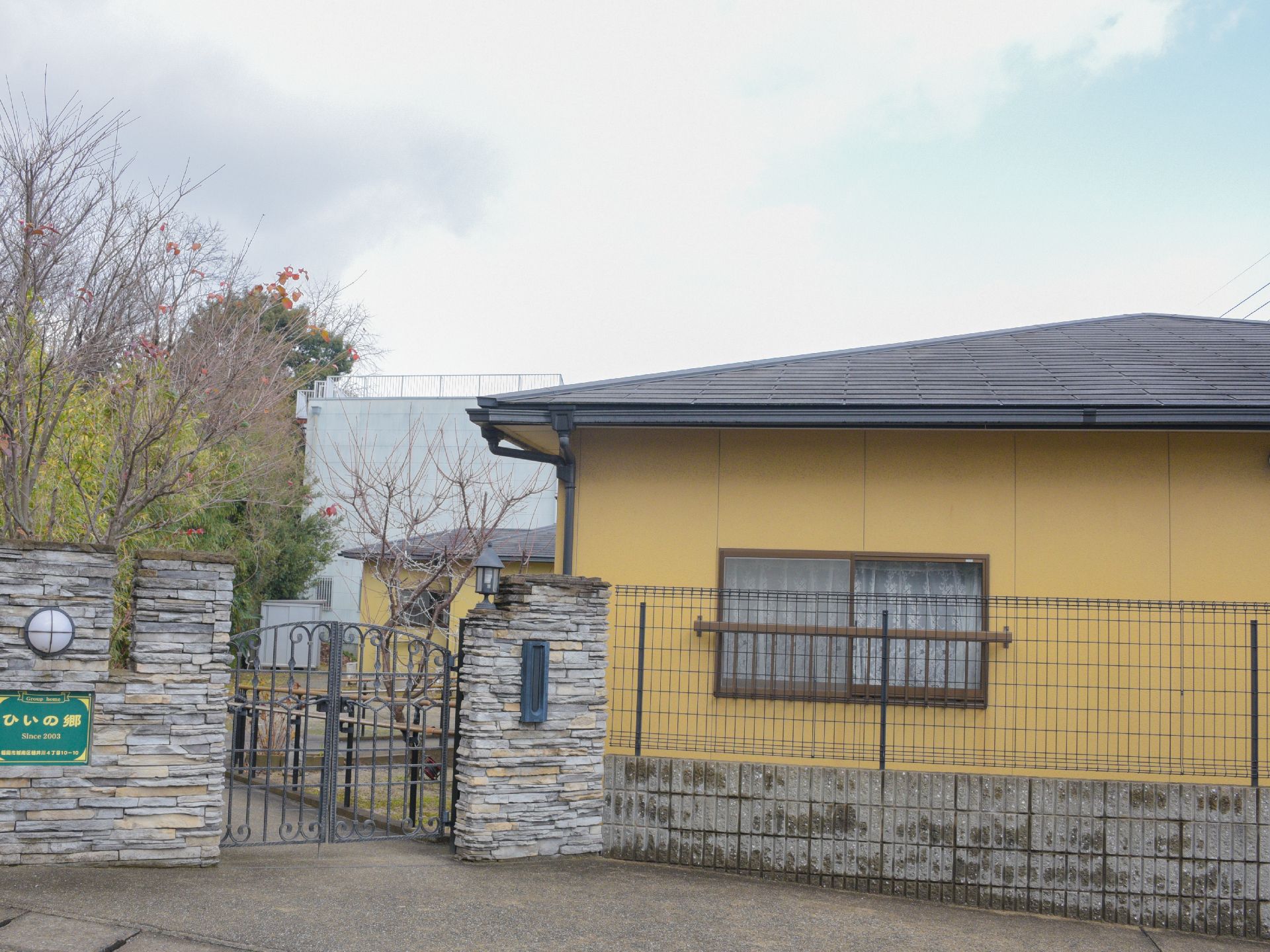 高齢者が暮らす居住施設の建物は、平屋建てとなっている。また入り口の門には看板があり、照明器具と郵便受けが設置されている。
