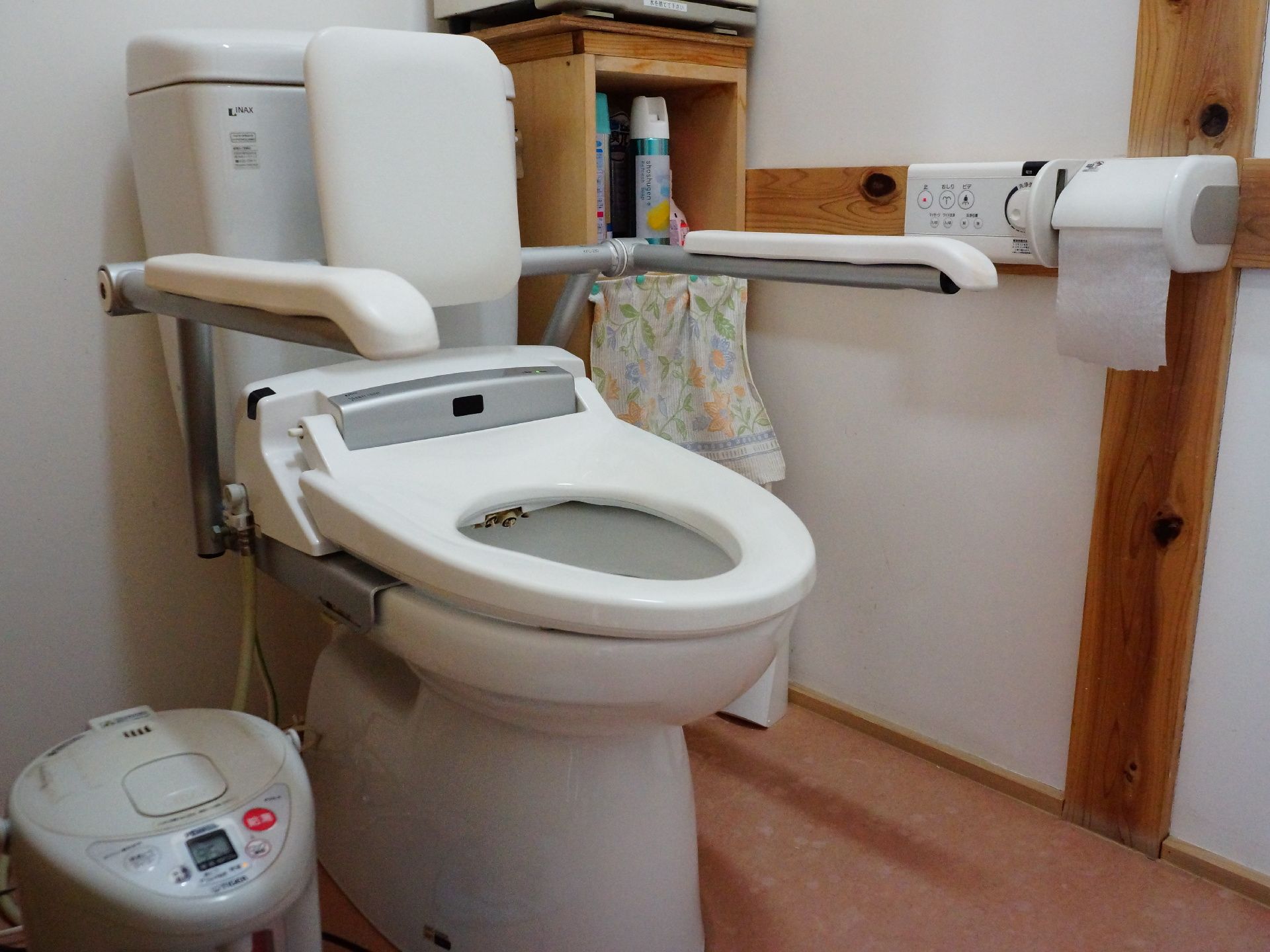 便器には背もたれと肘掛けがついている。その横には湯沸かし器がある。棚にはスプレー缶が置いてあり、その下段には小さなカーテンがかけられている。