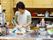 台所で女性が1人で調理をしている最中である。まな板に包丁、食器や食材がある。その後ろにはケトルや給湯器や鍋がある。