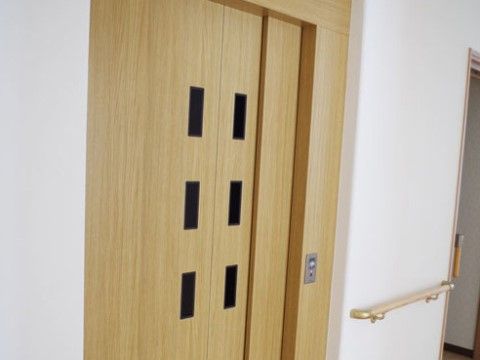 扉が木目調のエレベーターが１基設置されており、どなた様も利用できる。横の壁には、水平型手すりが取り付けられている。