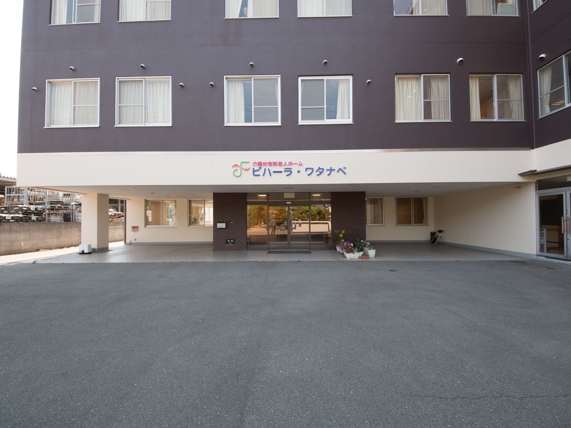 建物玄関の真上に、施設名が表示されている。出入口はガラスの自動ドアになっており、横に置かれたプランターには、花が植えられている。