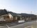 森林を背景にした平屋建ての和風の建物が建っている。建物の前にフェンスが設置され、アスファルトで整備された駐車場がある。