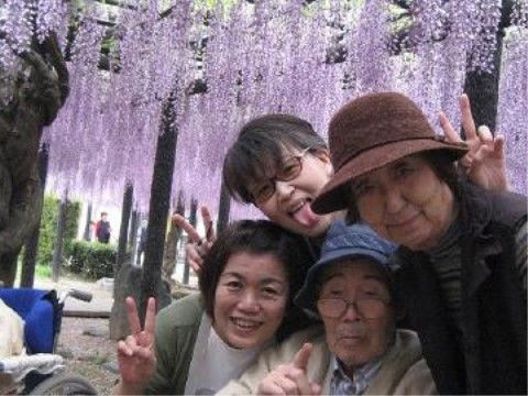 紫の長い藤の花が咲いた藤棚の前で笑っている入居者様